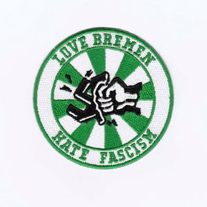 Könnte beinhalten: Runder Aufnäher mit grün-weißem Design. Der Text "LOVE BREMEN" ist oben in einem Bogen angeordnet, darunter "HATE FASCISM". In der Mitte befindet sich eine schwarz-weiße Grafik einer Faust, die eine Waffe umklammert.