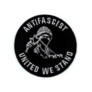 Könnte beinhalten: Schwarz-weiß gestickter Aufnäher mit einer vermummten Figur, die eine Waffe hält. Der Aufnäher ist rund und hat den Text "Anti-Faschist Vereint Stehen Wir" am Rand.