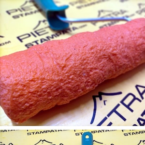 May include: A close-up of a textured orange paint roller with a blue handle. The roller is on a yellow surface with the text "PIETRA STAMPATA.COM" repeated.