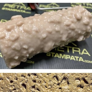 May include: A close-up of a textured paint roller with a pattern of small bumps. The roller is made of a light brown material and is designed to create a stone-like effect on walls. The roller is labeled "PIETRA STAMPATA" in black text.