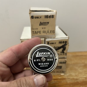 May include: A silver metal tape measure with a black face. The face reads "LUFKIN" in a white circle with a red "R" in the upper right corner. Below the circle, it reads "8 FT. 1688 WIZARD JUNIOR".