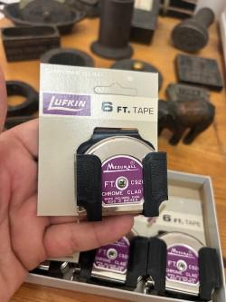 K&ouml;nnte beinhalten: Ein Lufkin 6 FT. (ca. 183 cm) Ma&szlig;band in einem schwarzen Halter. Das Ma&szlig;band hat eine verchromte Oberfl&auml;che und befindet sich in einer lila-wei&szlig;en Verpackung. Die Verpackung wird von einer Person gehalten. Weitere Werkzeuge sind im Hintergrund.