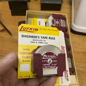 May include: A red and silver Lufkin engineer's tape measure with a chrome clad blade. The tape measure is graduated to 10ths and 100ths of a foot and is 2 feet long. The packaging says "See back for features".