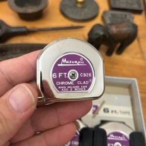 K&ouml;nnte beinhalten: Ein verchromtes Ma&szlig;band mit einem lila Etikett mit der Aufschrift "Mezurall 6 ft. C926". Das Ma&szlig;band wird in einer Hand gehalten, mit anderen Werkzeugen und Objekten im Hintergrund.