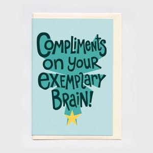 May include: A light blue greeting card featuring dark teal hand-lettered text that reads Compliments on your exemplary brain! The text is layered over a teal starburst shape with a small yellow star icon positioned at the bottom centre of the card.