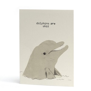 May include: A rectangular greeting card with a light beige background featuring a hand-drawn illustration of a grey dolphin head emerging from water. The text "dolphins are shit" is printed in a simple black sans-serif font above the dolphin.
