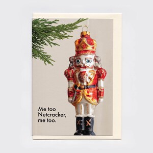 May include: A greeting card featuring a glass nutcracker ornament hanging from a green pine branch. The nutcracker wears a red and gold uniform with a crown. The text on the card reads, "Me too Nutcracker, me too." The design is set against a light background.