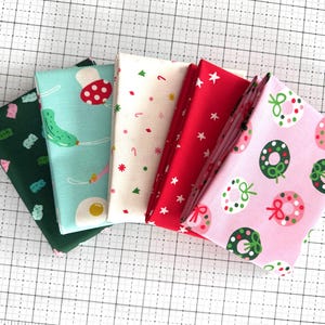 May include: Five folded fabric squares featuring various patterns: a dark green with gummy bears, a light blue with mushrooms and eggs, a white with candy canes and stars, a solid red with white stars, and a light pink with green and red wreaths.