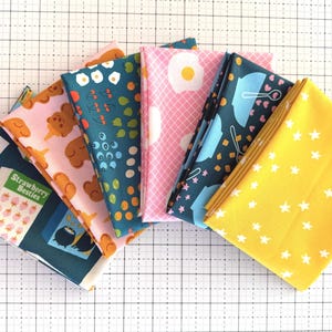 May include: A fan of folded fabric squares in various patterns and colors. Designs include strawberries, teddy bears, blueberries, eggs, kitchen utensils, and white stars on a yellow background. The fabrics are arranged on a white grid.