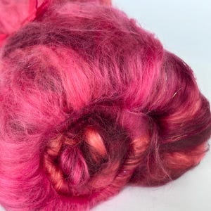 Peut inclure: Gros plan sur une fibre rose vibrante et multicolore. La fibre est un mélange de rose vif, de bordeaux profond et de nuances plus claires, créant un aspect texturé et superposé. Un petit ruban rose est visible.
