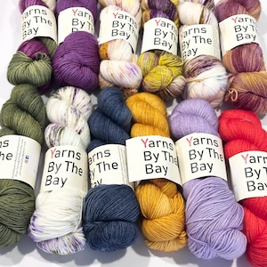 May include: Twelve skeins of hand-dyed yarn arranged in two rows. Each skein features a white paper label printed with the text Yarns By The Bay. The collection includes various solid and variegated colors such as olive green, purple, mustard yellow, navy blue, lavender, bright red, and cream with speckles.