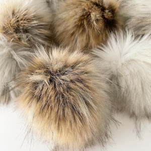 May include: Five fluffy pom poms in various shades of brown and white. The pom poms are made of faux fur and are soft and plush.