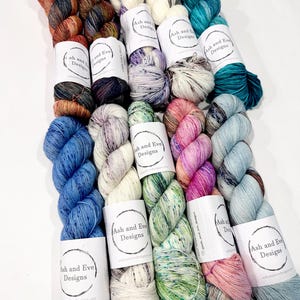 May include: A collection of twelve skeins of yarn arranged in rows, each featuring a white paper label with the text Ash and Eve Designs. The yarn displays a variety of variegated colours including shades of blue, green, pink, orange, and cream.