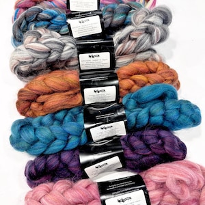 May include: A collection of seven braided wool roving bundles in various colours including pink, purple, teal, orange, grey, and multi-coloured blends. Each bundle is secured with a black paper label containing product information and text.
