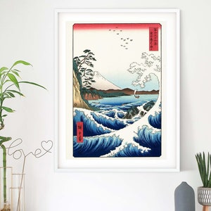 May include: A framed print of a Japanese woodblock print depicting a large wave crashing against a rocky shore. The wave is white with blue and green accents. A small boat with a sail is in the distance. The print is set against a white wall with a green plant in a vase to the left and a gray diffuser to the right.