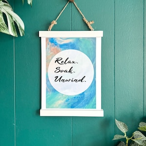 May include: White framed print with a blue and pink abstract background. The print has a white circle with the text "Relax. Soak. Unwind." in black.