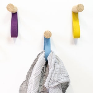 May include: Three wooden wall hooks with fabric loops in purple, yellow, and blue. A light gray cloth is hanging on the blue hook.