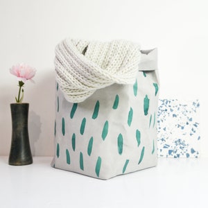 以下が含まれることがあります： 緑色の抽象的な模様がついた白い紙袋。袋には白いニットのストールが入っています。