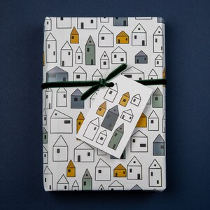 Op de afbeelding: Wit cadeaupapier met een herhalend patroon van kleine huisjes in verschillende kleuren. Een groen lint is om het pakket gebonden met een wit cadeaukaartje dat een patroon van kleine huisjes in verschillende kleuren heeft.