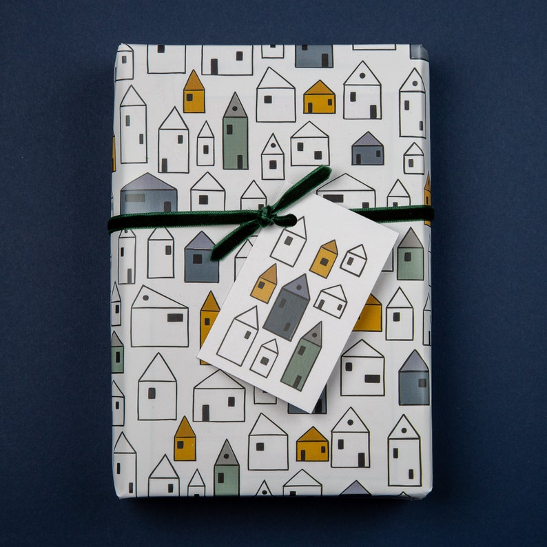 Op de afbeelding: Wit cadeaupapier met een herhalend patroon van kleine huisjes in verschillende kleuren. Een groen lint is om het pakket gebonden met een wit cadeaukaartje dat een patroon van kleine huisjes in verschillende kleuren heeft.