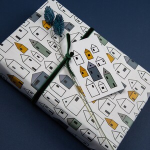 Op de afbeelding: Wit cadeaupapier met een herhalend patroon van kleine huisjes in zwart, geel en groen. Een groen fluwelen lint is om het cadeau gebonden met een wit cadeaukaartje dat een patroon van kleine huisjes in zwart, geel en blauw heeft.