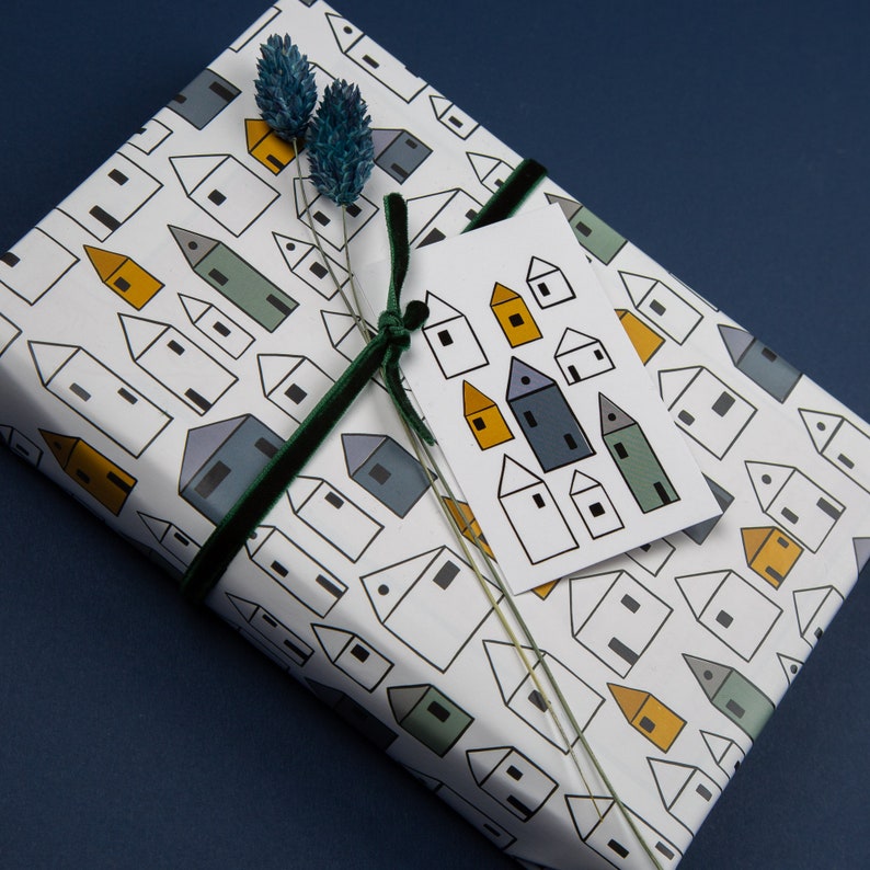 Op de afbeelding: Wit cadeaupapier met een herhalend patroon van kleine huisjes in zwart, geel en groen. Een groen fluwelen lint is om het cadeau gebonden met een wit cadeaukaartje dat een patroon van kleine huisjes in zwart, geel en blauw heeft.