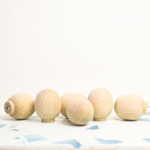 Puede incluir: Seis pomos de madera sin terminar en tono marrón claro. Los pomos tienen forma redondeada, con diferentes diseños. Algunos tienen base plana, otros una pequeña extensión cilíndrica. Están dispuestos sobre una superficie blanca y azul.