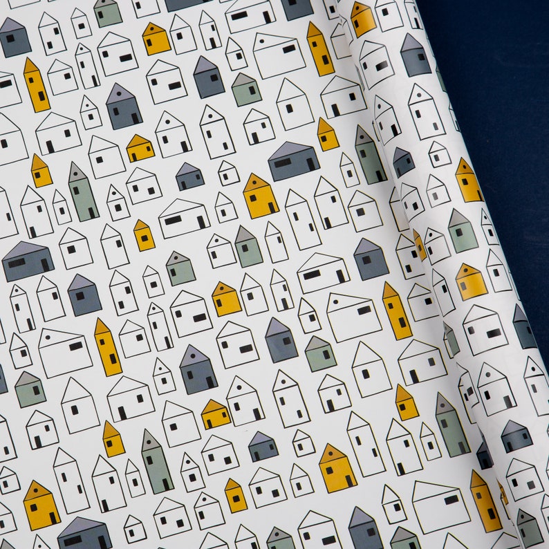 Op de afbeelding: Wit inpakpapier met een herhalend patroon van kleine huisjes in verschillende tinten blauw, grijs en geel. De huisjes zijn zwart omlijnd en hebben een eenvoudig, modern ontwerp.