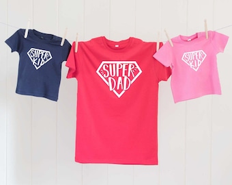 May include: Three t-shirts hanging on a clothesline. The center shirt is red with white text that says "Super Dad" in a superhero logo. The shirt on the left is navy blue with white text that says "Super Kid". The shirt on the right is pink with white text that says "Super Kid".