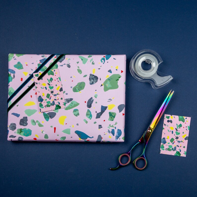 May include: Pink wrapping paper with a terrazzo pattern. A black ribbon, a clear tape dispenser, a pair of rainbow scissors, and two gift tags are also included.