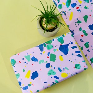 May include: Pink wrapping paper with a terrazzo pattern in blue, green, yellow, and red. The paper is on a yellow surface with a small potted plant.