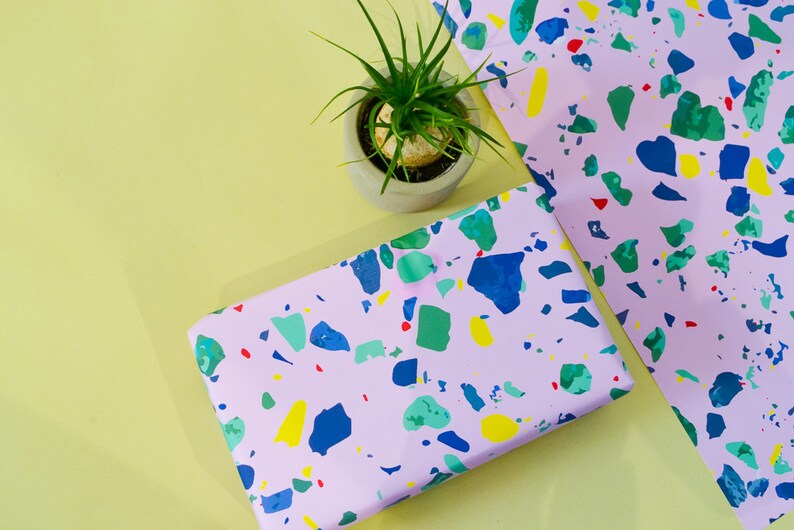 May include: Pink wrapping paper with a terrazzo pattern in blue, green, yellow, and red. The paper is on a yellow surface with a small potted plant.