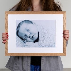 May include: A framed photograph of a sleeping baby wrapped in a white blanket. The photo is in a light wood frame with a white mount.