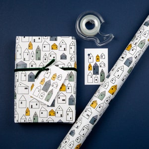 Op de afbeelding: Wit cadeaupapier met een herhalend patroon van kleine huisjes in zwart, blauw en geel. Een bijpassend cadeaukaartje met een vergelijkbaar patroon is inbegrepen. Een rol doorzichtige tape is ook afgebeeld.