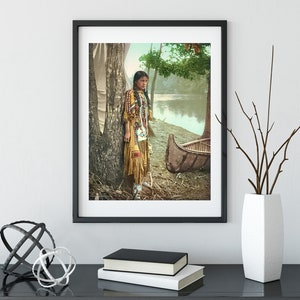 May include: A vintage photograph of a Native American woman standing by a tree and a canoe. She is wearing a traditional beaded dress and has long dark hair. The photo is in color and has a faded look.