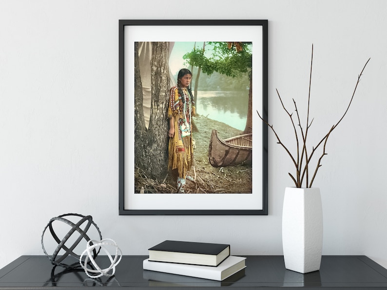 May include: A vintage photograph of a Native American woman standing by a tree and a canoe. She is wearing a traditional beaded dress and has long dark hair. The photo is in color and has a faded look.