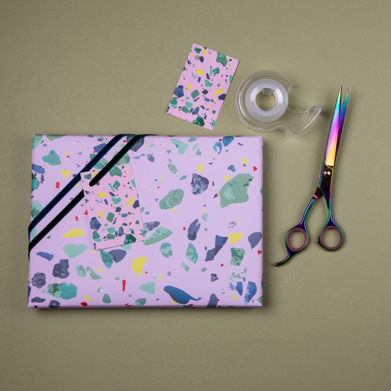 May include: A pink and white terrazzo patterned gift wrap with a black ribbon, a matching gift tag, a roll of clear tape, and a pair of rainbow-colored scissors.