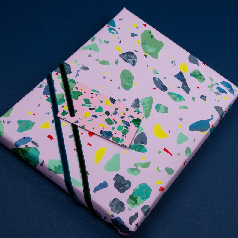 May include: Pink gift wrap with a terrazzo pattern in shades of green, blue, yellow, and red. A pink gift tag with a matching terrazzo pattern is attached to the wrapping paper with black ribbon.