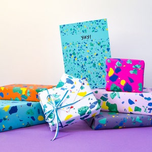 May include: A stack of colorful gift boxes wrapped in terrazzo patterned paper. The boxes are arranged in a pyramid shape with a card on top that says "yay!"