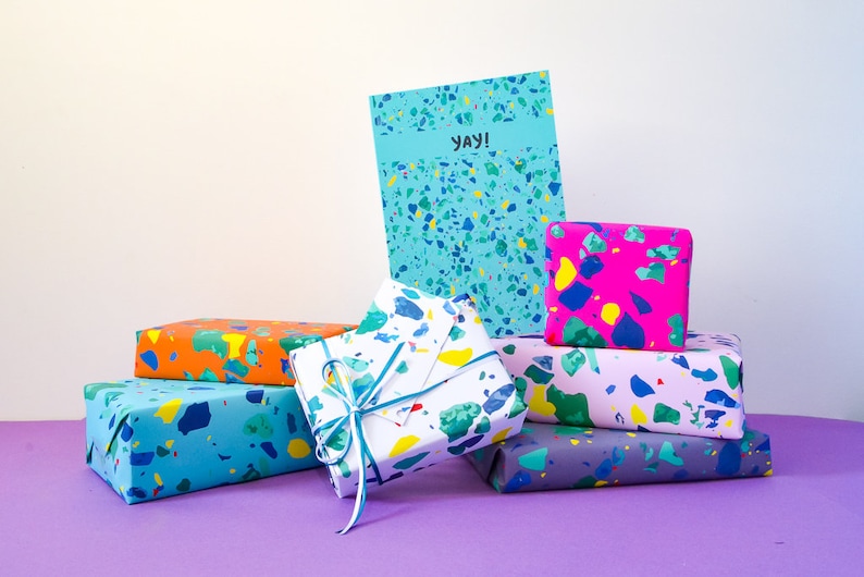 May include: A stack of colorful gift boxes wrapped in terrazzo patterned paper. The boxes are arranged in a pyramid shape with a card on top that says "yay!"