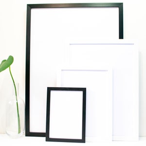 May include: Three picture frames of different sizes, one black and two white, are arranged on a white surface. The largest frame is black and the two smaller frames are white. A single green leaf is in a clear glass vase.