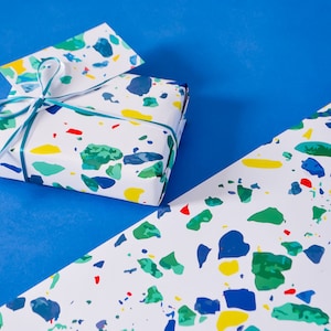 May include: A gift wrapped in white paper with a colorful terrazzo pattern. The gift is tied with a teal ribbon and a white ribbon. The gift is sitting on a blue surface.