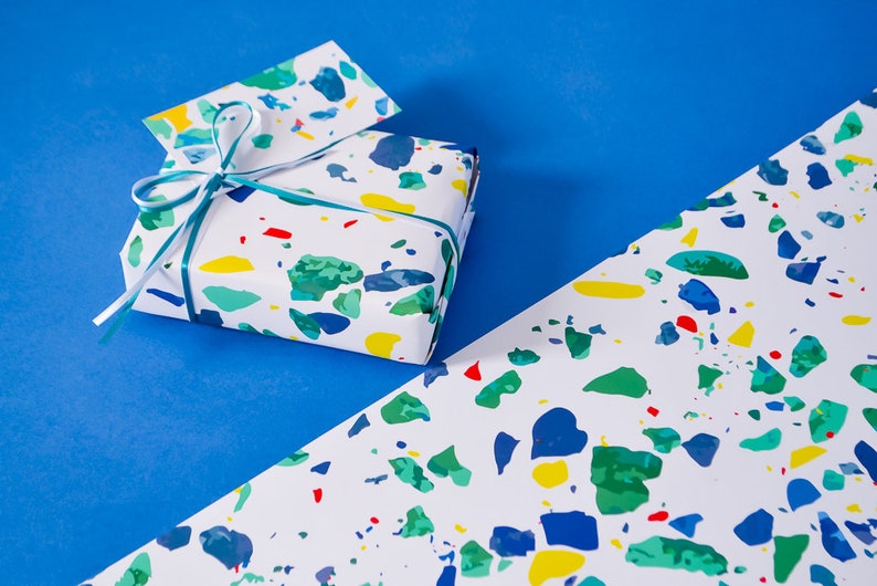 May include: A gift wrapped in white paper with a colorful terrazzo pattern. The gift is tied with a teal ribbon and a white ribbon. The gift is sitting on a blue surface.