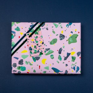 May include: Pink gift wrap with a terrazzo pattern in shades of green, yellow, and black. A small pink gift tag with a similar pattern is attached with a black ribbon.