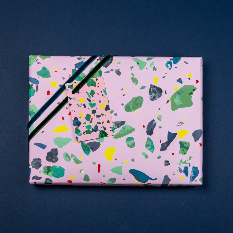 May include: Pink gift wrap with a terrazzo pattern in shades of green, yellow, and black. A small pink gift tag with a similar pattern is attached with a black ribbon.