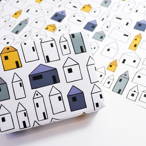 Op de afbeelding: Wit inpakpapier met een herhalend patroon van kleurrijke huizen. De huizen zijn in tinten blauw, geel en grijs. Het papier is gevouwen en klaar om te worden gebruikt voor het inpakken van cadeaus.