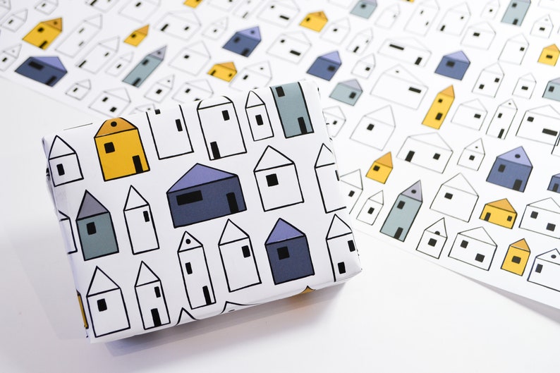 Op de afbeelding: Wit inpakpapier met een herhalend patroon van kleurrijke huizen. De huizen zijn in tinten blauw, geel en grijs. Het papier is gevouwen en klaar om te worden gebruikt voor het inpakken van cadeaus.