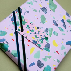 May include: Pink wrapping paper with a colorful terrazzo pattern. The paper is decorated with a pink gift tag with a matching terrazzo pattern. The paper is tied with a black and green velvet ribbon.