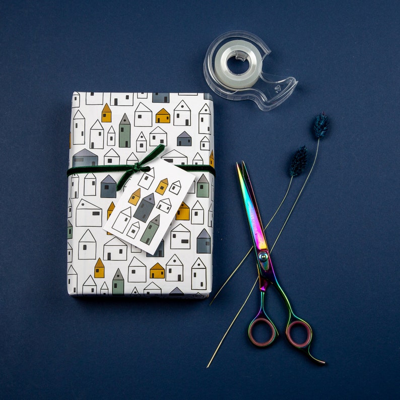 Op de afbeelding: Wit cadeaupapier met een patroon van kleine huisjes in verschillende kleuren. Een groen lint is om het pakket gebonden. Een doorzichtige tape dispenser en regenboogkleurige schaar zijn ook op de afbeelding te zien.