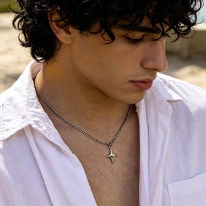 Può includere: Una collana in metallo color argento con una sottile catena a maglie e un ciondolo a forma di stella a quattro punte. Il ciondolo presenta una finitura liscia e lucida con punte allungate e affusolate. La collana è indossata su una camicia bianca.
